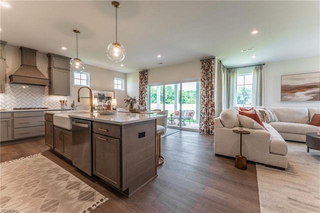 kitchen with hardwood / wood-style floors, custom exhaust hood, hanging light fixtures, an island with sink, and appliances with stainless steel finishes