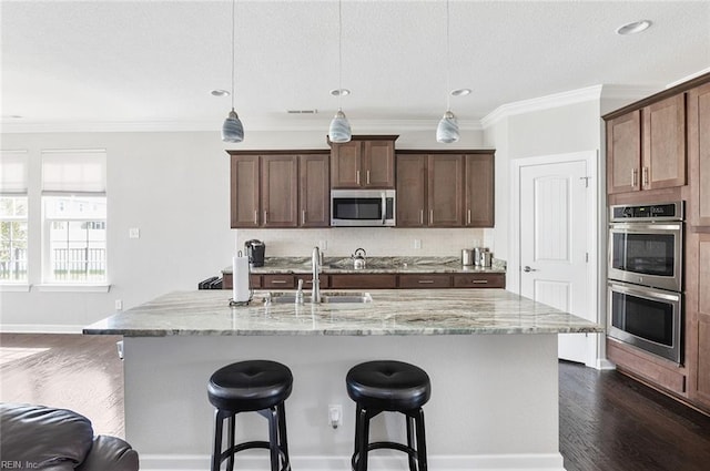 kitchen with hanging light fixtures, stainless steel appliances, light stone counters, dark hardwood / wood-style floors, and an island with sink