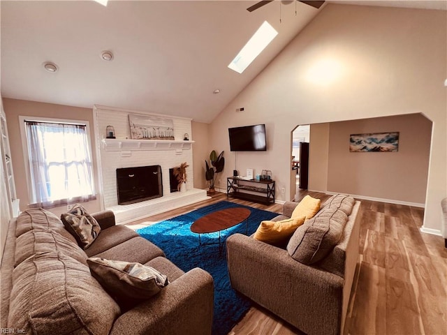 living room with light wood-type flooring, a skylight, a brick fireplace, ceiling fan, and high vaulted ceiling