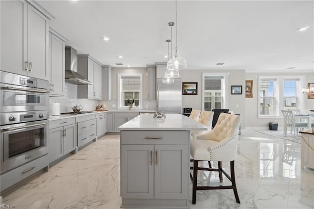 kitchen featuring a center island, wall chimney exhaust hood, gray cabinets, appliances with stainless steel finishes, and decorative light fixtures