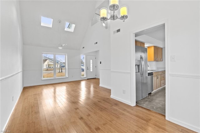 unfurnished living room with ceiling fan with notable chandelier, high vaulted ceiling, a skylight, and light hardwood / wood-style flooring