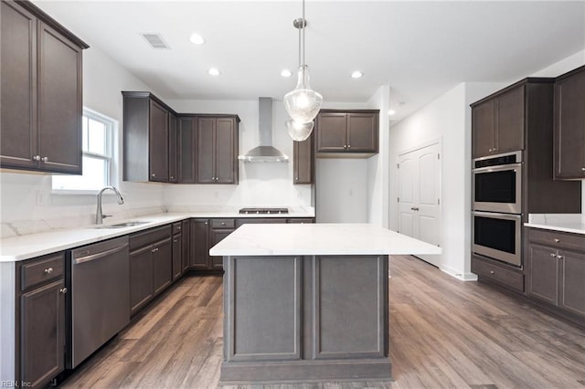 kitchen with a center island, sink, hanging light fixtures, stainless steel appliances, and wall chimney range hood