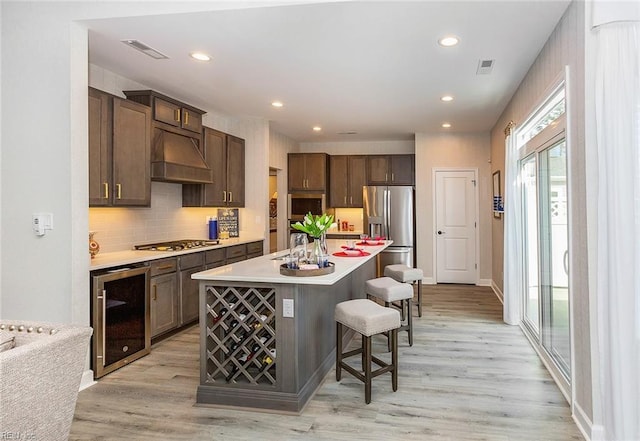 kitchen featuring appliances with stainless steel finishes, a kitchen breakfast bar, custom range hood, beverage cooler, and a center island with sink