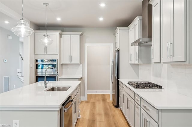 kitchen with decorative light fixtures, wall chimney range hood, a kitchen island with sink, and appliances with stainless steel finishes