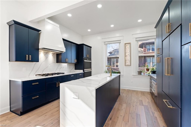 kitchen with backsplash, premium range hood, blue cabinets, light hardwood / wood-style flooring, and an island with sink