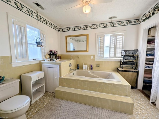 bathroom featuring tile walls, ceiling fan, toilet, tiled tub, and a textured ceiling