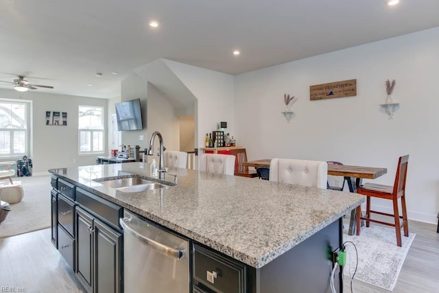 kitchen featuring a kitchen island with sink, sink, stainless steel dishwasher, ceiling fan, and light stone countertops