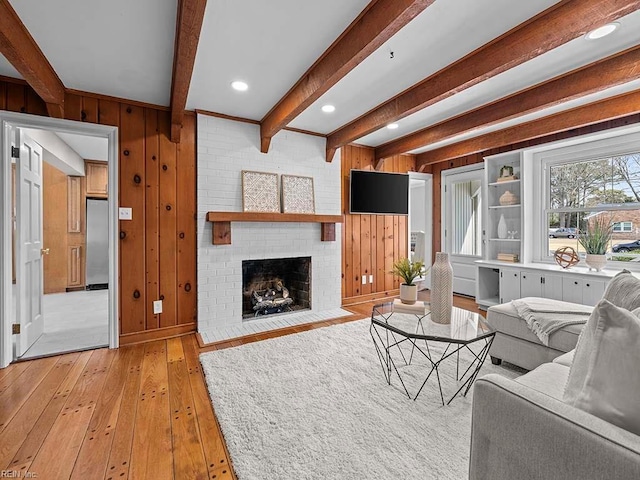 living room featuring a brick fireplace, light wood-type flooring, wooden walls, and beamed ceiling