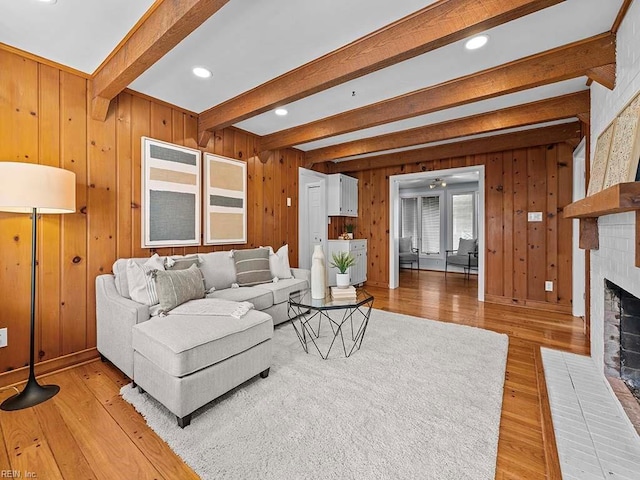 living room featuring wooden walls, a fireplace, beamed ceiling, light wood-type flooring, and ceiling fan