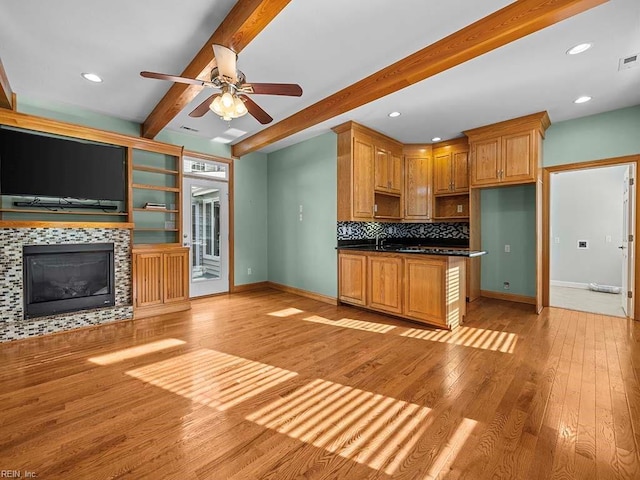 kitchen featuring light hardwood / wood-style floors, a tiled fireplace, beamed ceiling, ceiling fan, and backsplash