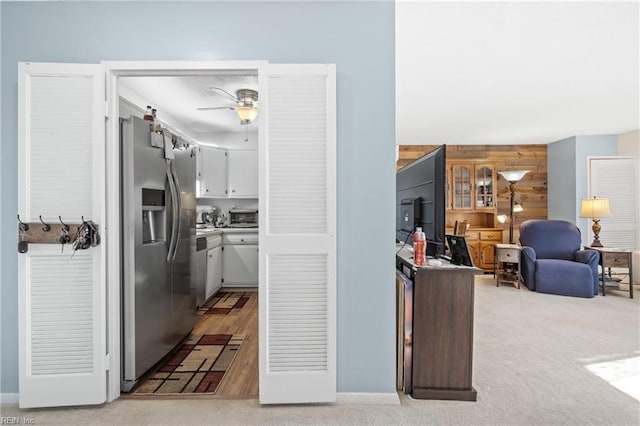 kitchen featuring wood walls, light colored carpet, ceiling fan, stainless steel refrigerator with ice dispenser, and white cabinets