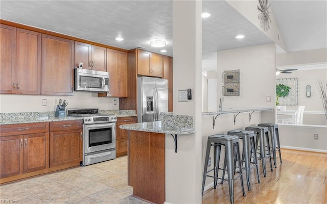 kitchen with kitchen peninsula, light stone countertops, stainless steel appliances, a kitchen bar, and ceiling fan