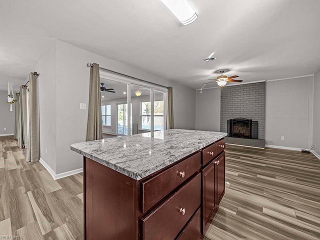 kitchen with a center island, light wood-type flooring, ceiling fan, a brick fireplace, and light stone countertops