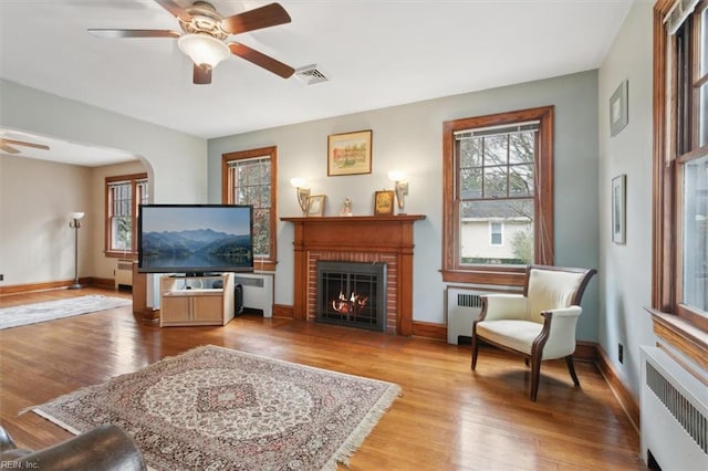 living room featuring a brick fireplace, radiator heating unit, light wood-type flooring, and ceiling fan