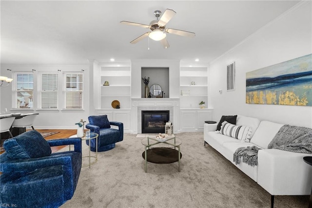 carpeted living room featuring crown molding, a tiled fireplace, built in features, and ceiling fan with notable chandelier