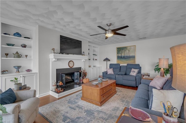 living room with ceiling fan, built in shelves, light hardwood / wood-style flooring, and a textured ceiling