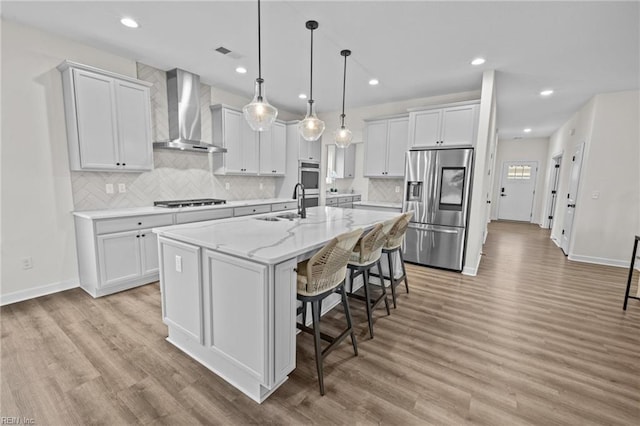 kitchen with wall chimney range hood, a center island with sink, white cabinets, and appliances with stainless steel finishes