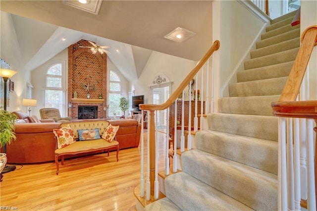 staircase with ceiling fan, hardwood / wood-style floors, a brick fireplace, and high vaulted ceiling