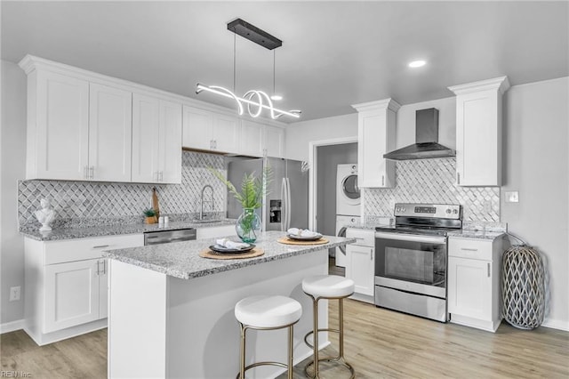 kitchen featuring stacked washer and dryer, wall chimney range hood, stainless steel appliances, white cabinets, and decorative light fixtures