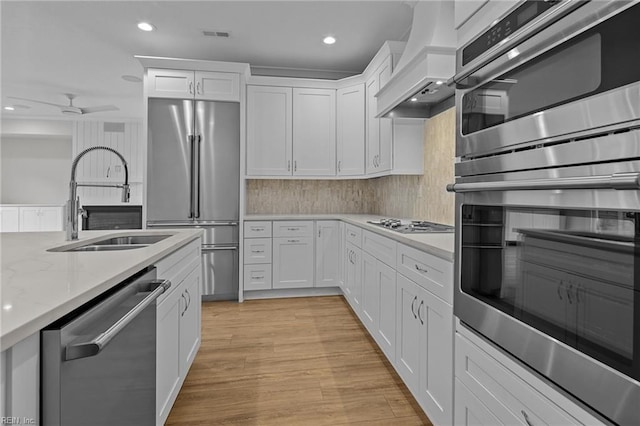 kitchen with light wood-style flooring, appliances with stainless steel finishes, custom exhaust hood, white cabinetry, and a sink