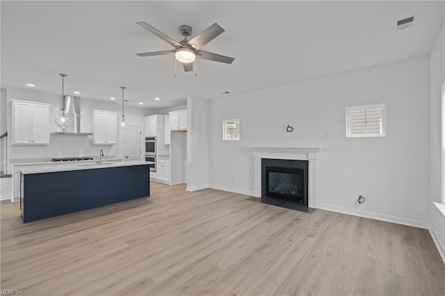 kitchen featuring decorative light fixtures, white cabinetry, wall chimney range hood, a center island with sink, and light wood-type flooring