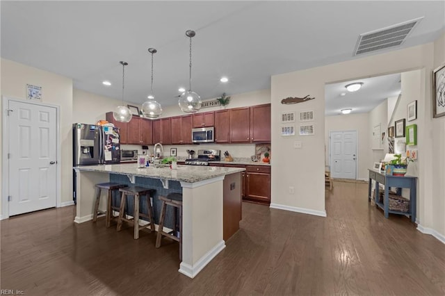 kitchen with dark wood-type flooring, decorative light fixtures, appliances with stainless steel finishes, an island with sink, and light stone countertops