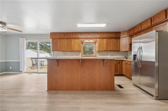 kitchen with stainless steel fridge, a kitchen breakfast bar, decorative backsplash, light stone countertops, and light wood-type flooring