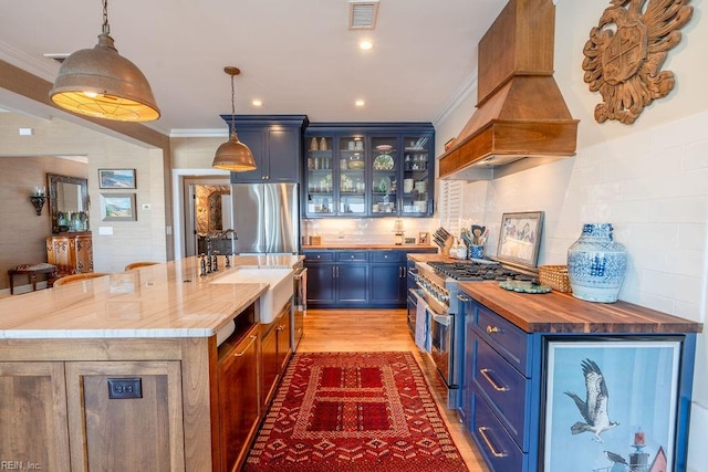 kitchen featuring blue cabinetry, appliances with stainless steel finishes, hanging light fixtures, light stone countertops, and an island with sink