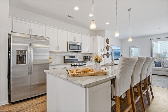 kitchen featuring white cabinetry, stainless steel appliances, light stone counters, an island with sink, and decorative light fixtures