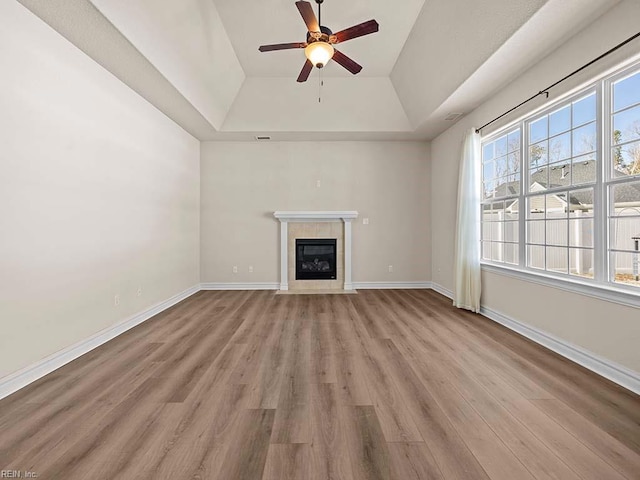 unfurnished living room featuring a tiled fireplace, a tray ceiling, ceiling fan, and light wood-type flooring