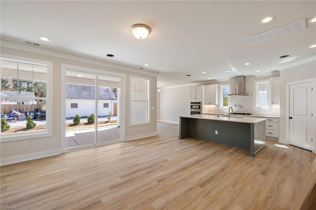 kitchen featuring white cabinetry, ornamental molding, a center island with sink, wall chimney range hood, and light wood-type flooring