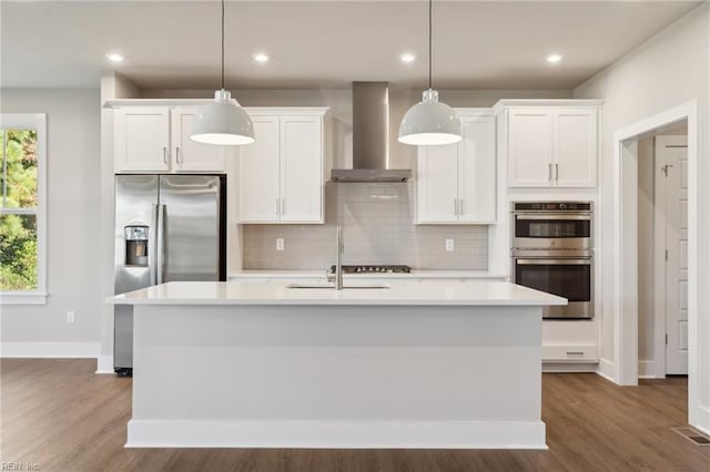 kitchen with appliances with stainless steel finishes, a kitchen island with sink, pendant lighting, and wall chimney exhaust hood