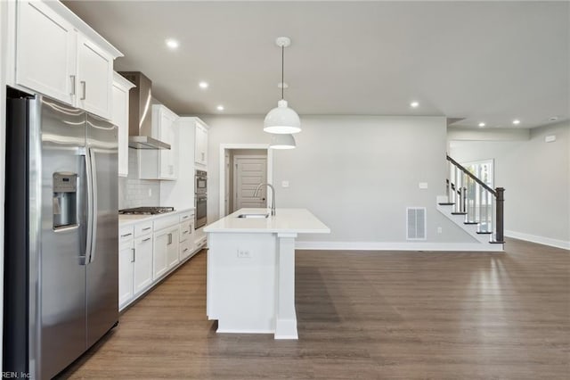 kitchen featuring appliances with stainless steel finishes, an island with sink, backsplash, hanging light fixtures, and wall chimney exhaust hood