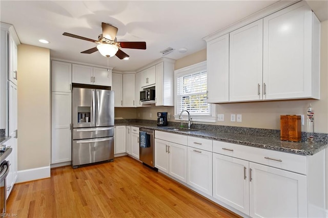 kitchen with appliances with stainless steel finishes, sink, white cabinets, dark stone counters, and light hardwood / wood-style flooring