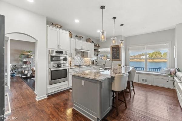 kitchen with a kitchen island with sink, double oven, a water view, light stone counters, and white cabinets