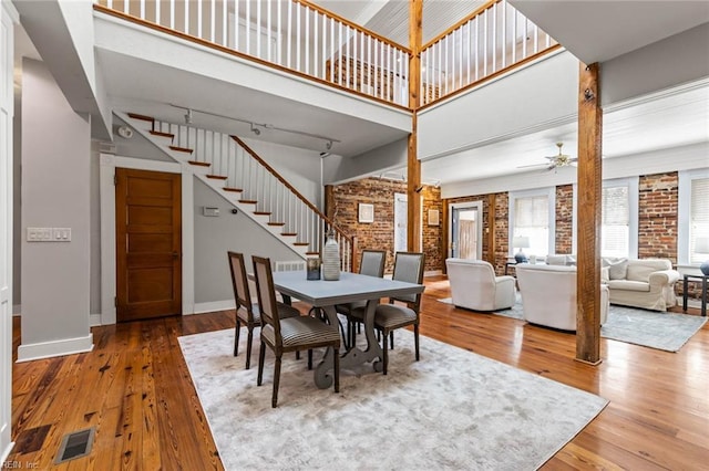 dining area featuring rail lighting, wood-type flooring, ceiling fan, brick wall, and a high ceiling