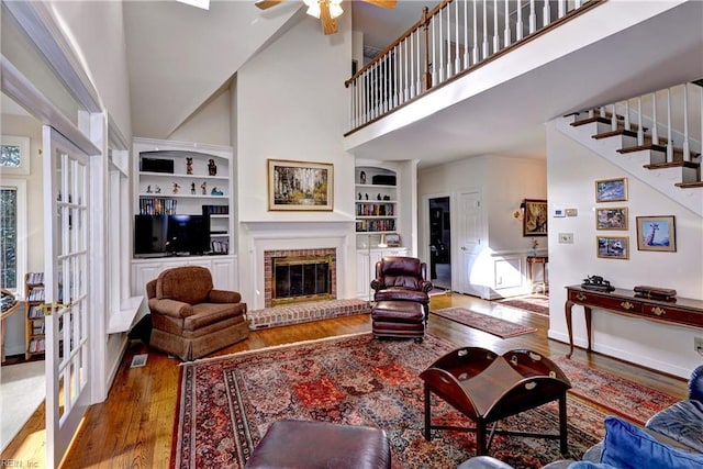 living room with wood-type flooring, built in features, ceiling fan, a fireplace, and a high ceiling