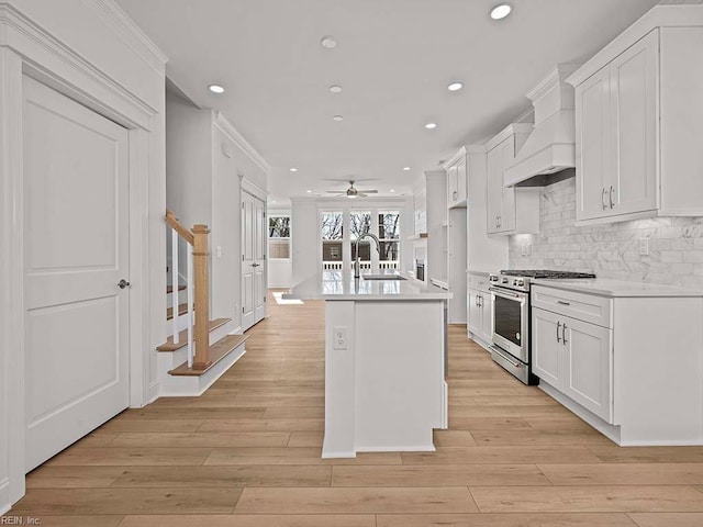 kitchen featuring premium range hood, an island with sink, sink, white cabinets, and stainless steel range