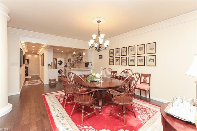 dining room with dark wood-type flooring, stacked washing maching and dryer, a notable chandelier, ornamental molding, and ornate columns