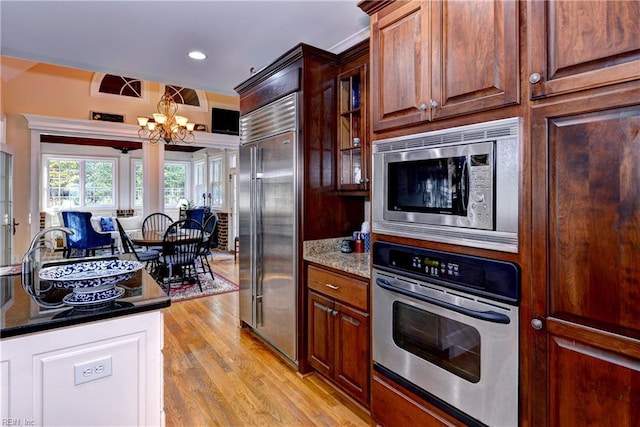 kitchen featuring sink, dark stone countertops, an inviting chandelier, built in appliances, and light wood-type flooring