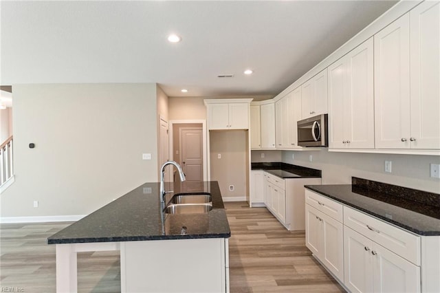 kitchen with sink, light wood-type flooring, dark stone counters, a kitchen island with sink, and white cabinets