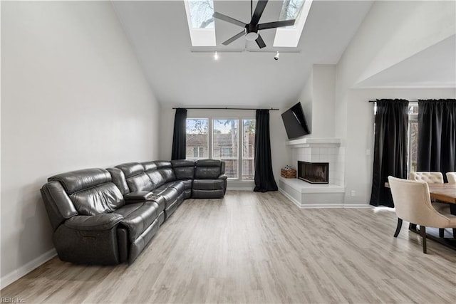 living room featuring high vaulted ceiling, a fireplace, a skylight, ceiling fan, and light hardwood / wood-style floors