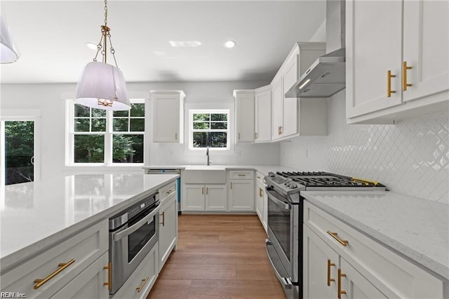 kitchen featuring wall chimney range hood, white cabinetry, stainless steel appliances, light stone countertops, and decorative light fixtures