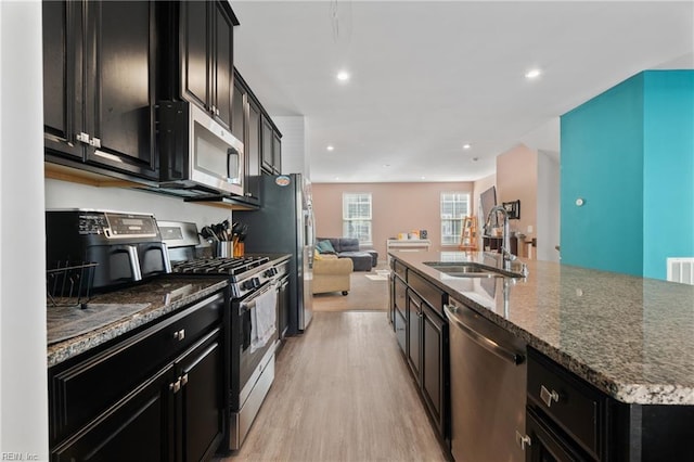 kitchen with sink, appliances with stainless steel finishes, dark stone countertops, light hardwood / wood-style floors, and an island with sink