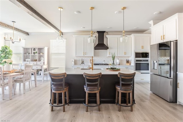 kitchen featuring appliances with stainless steel finishes, pendant lighting, white cabinets, a center island with sink, and wall chimney exhaust hood