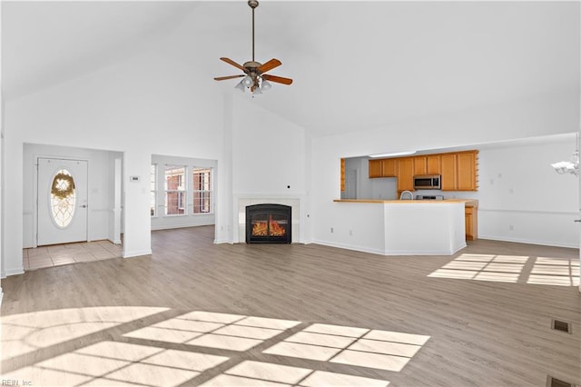 unfurnished living room with high vaulted ceiling, light wood-type flooring, ceiling fan with notable chandelier, and a fireplace