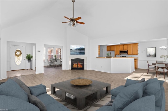 living room with a tiled fireplace, ceiling fan with notable chandelier, high vaulted ceiling, and light hardwood / wood-style flooring