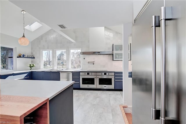 kitchen featuring wall chimney range hood, hanging light fixtures, lofted ceiling with skylight, stainless steel appliances, and decorative backsplash