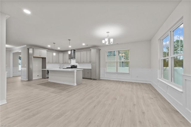 kitchen featuring wall chimney range hood, gray cabinets, hanging light fixtures, a center island with sink, and a chandelier