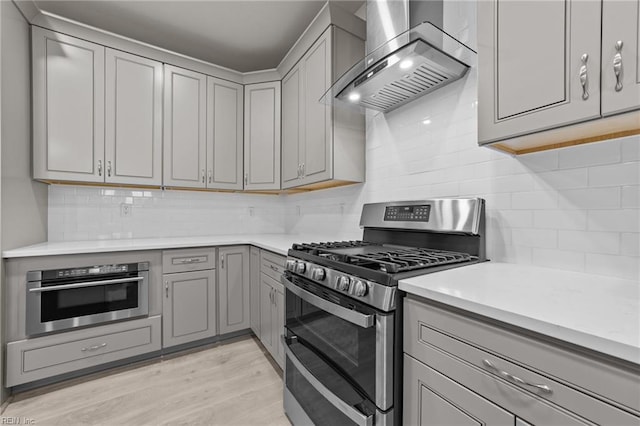 kitchen with ventilation hood, light wood-type flooring, appliances with stainless steel finishes, gray cabinets, and backsplash